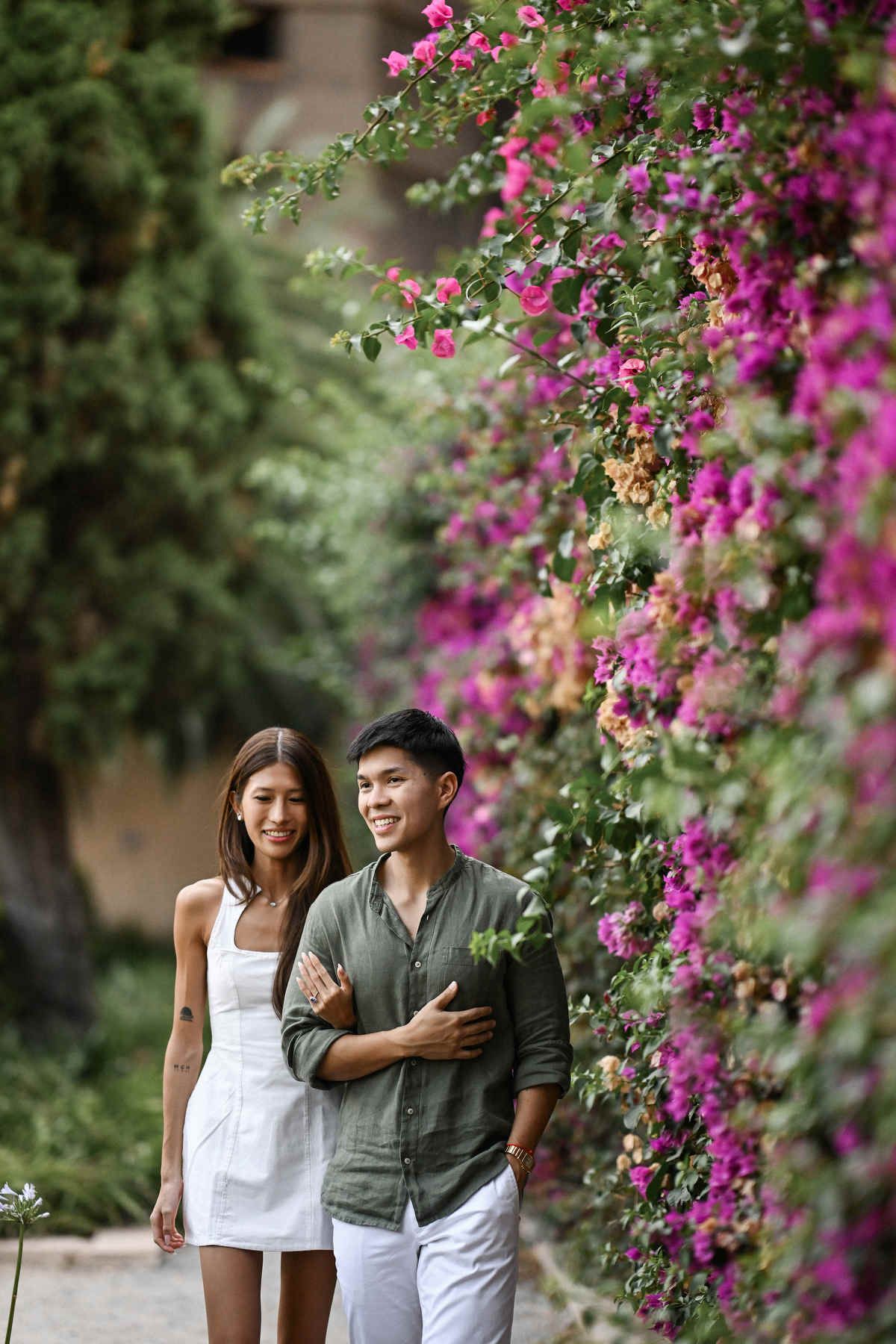 Engaged couple walking beside a stunning pink bougainvillea wall during their love story session in Monforte Gardens, Valencia.