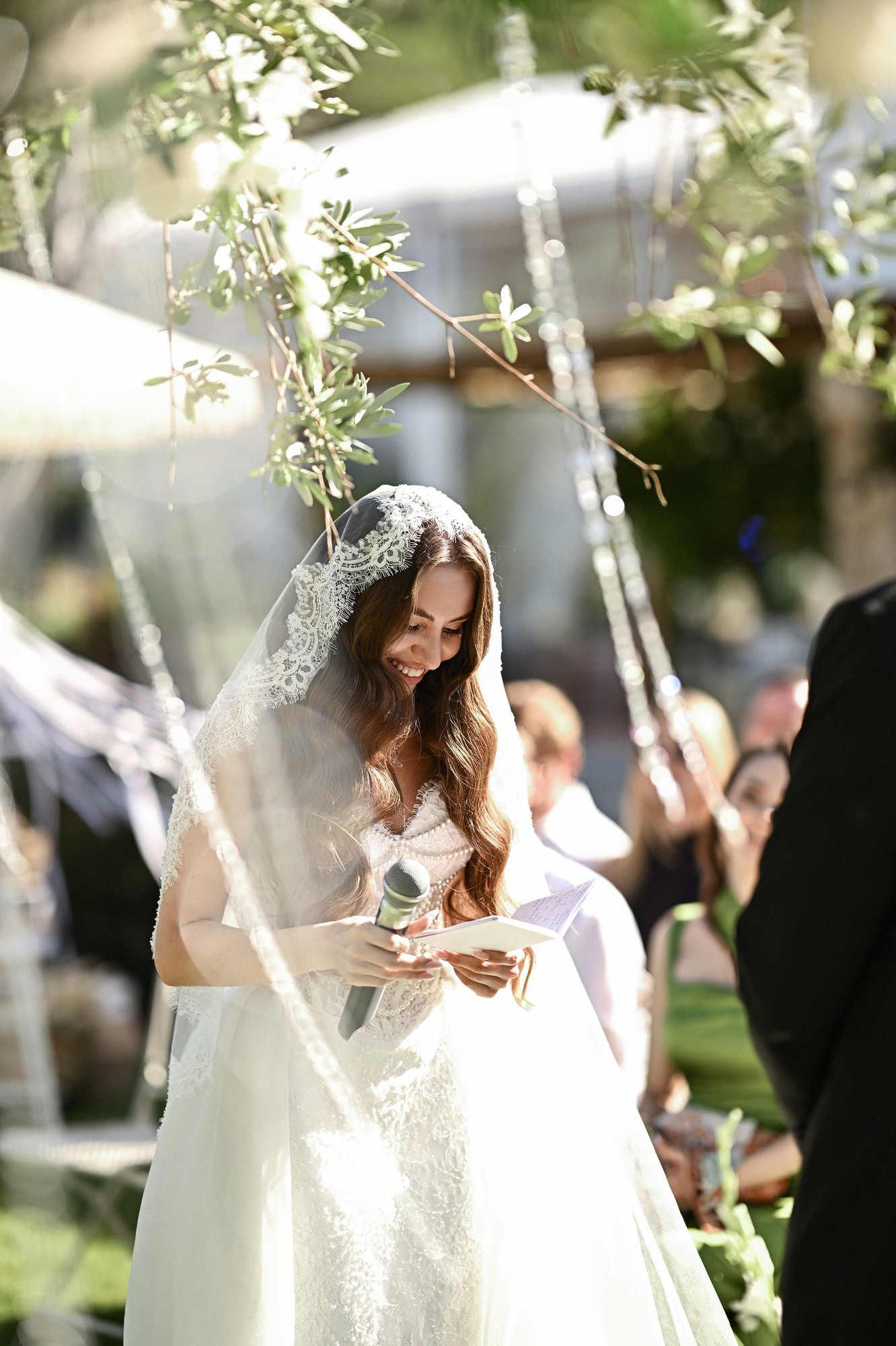 Smiling bride wearing a classic lace veil reading her vows during an outdoor winery wedding ceremony in Spain.