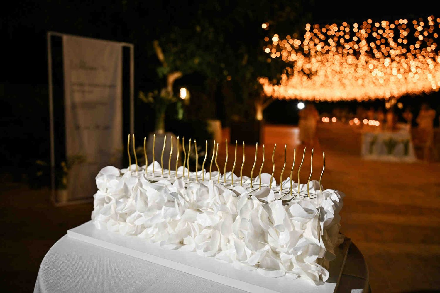Rectangle white textured wedding cake decorated with long golden candles at an evening reception.