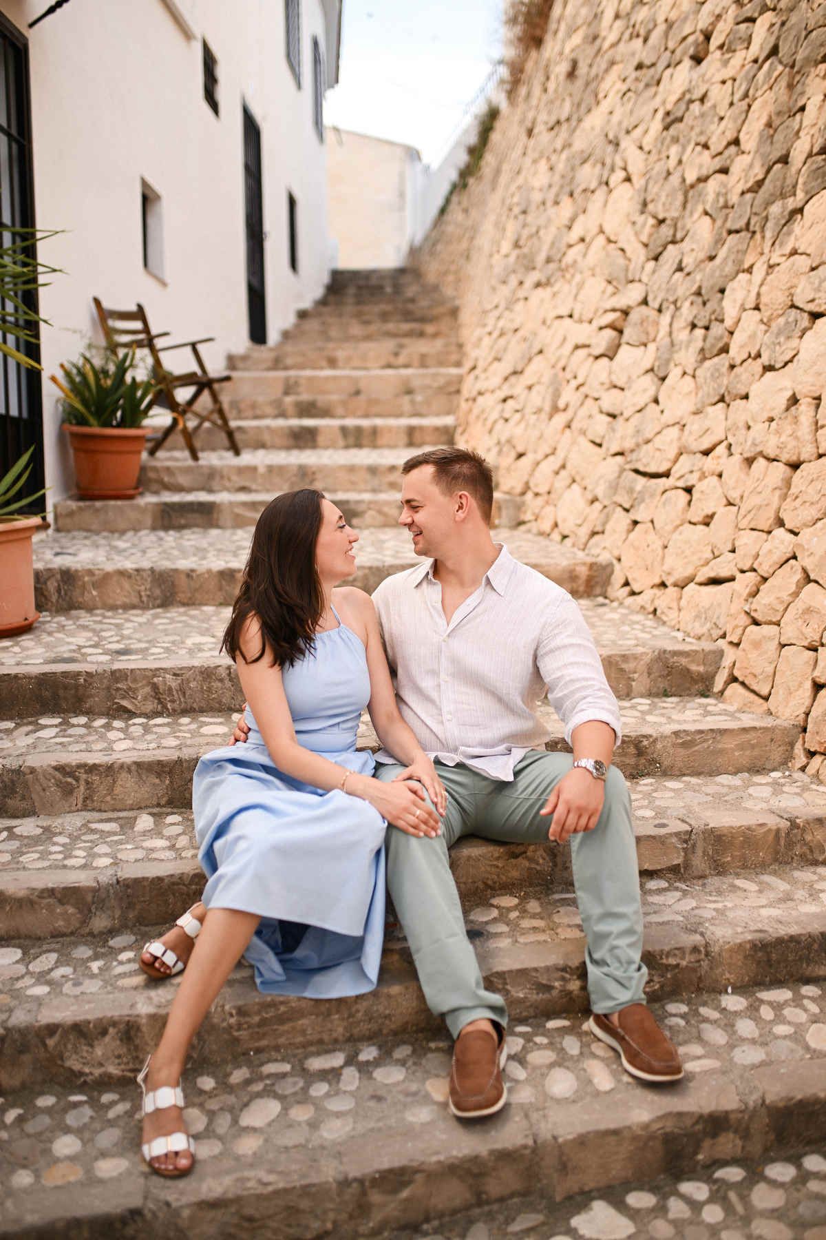 A newly engaged couple sits closely together on old stone stairs against a textured rock wall in Altea Old Town.