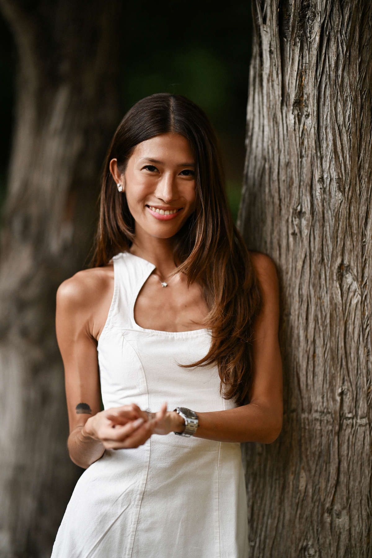 A smiling woman in a white dress leaning against a thick tree trunk to celebrate her surprise proposal in Monforte.