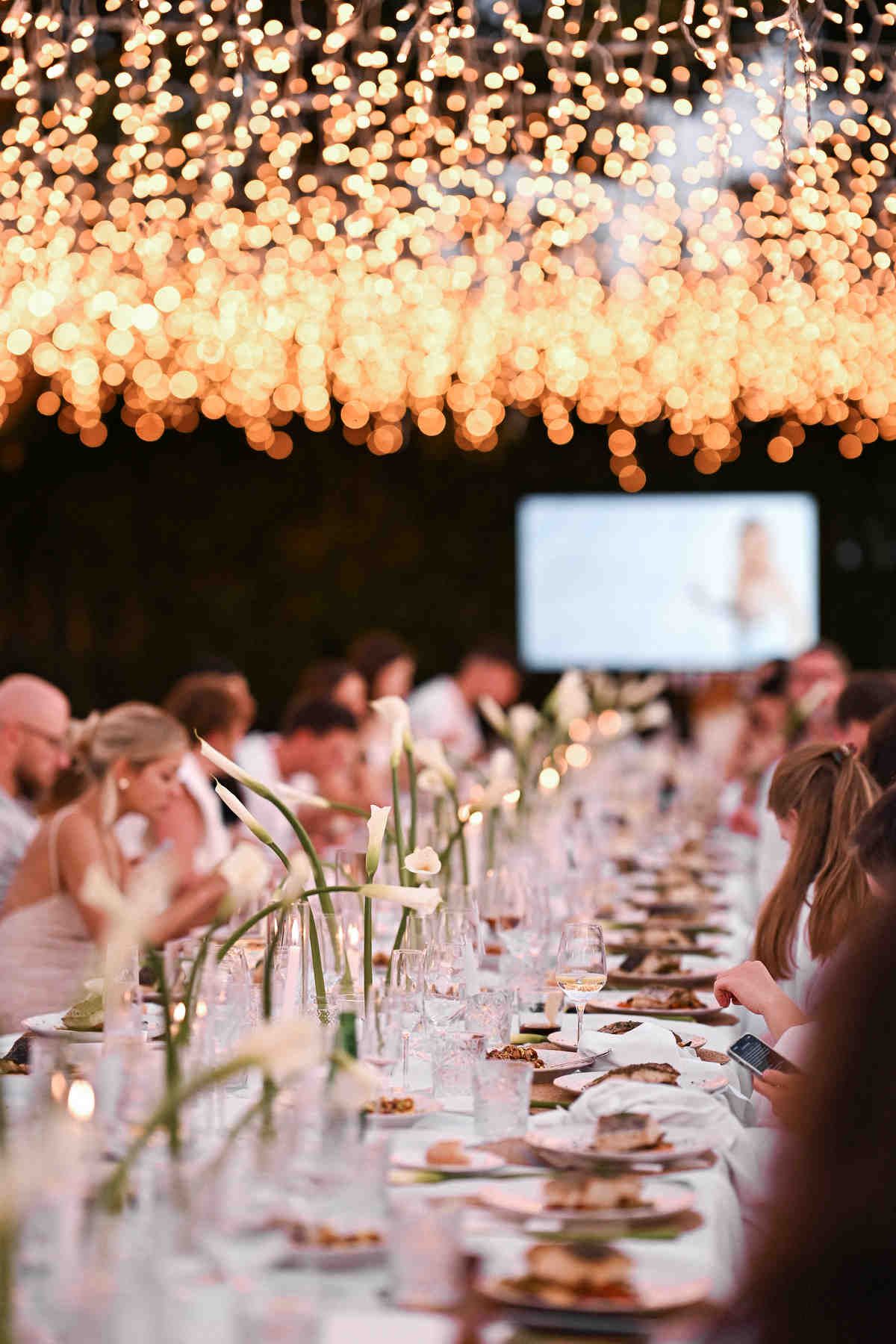Wedding reception table with guests dining under fairy lights with a large presentation screen in the background.