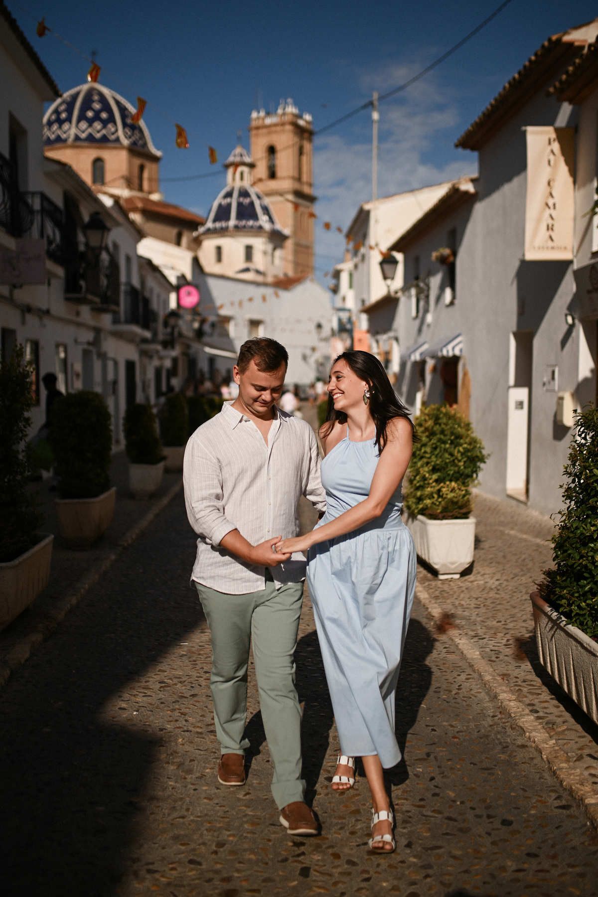 A smiling couple walks hand in hand down a narrow, sunlit whitewashed street during their Altea proposal session.