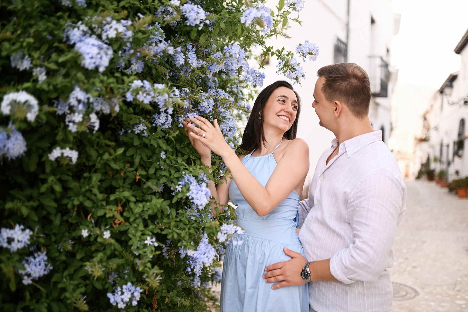 A proposal photographer in Altea captures a smiling couple by a blue flowering bush in a narrow whitewashed street.