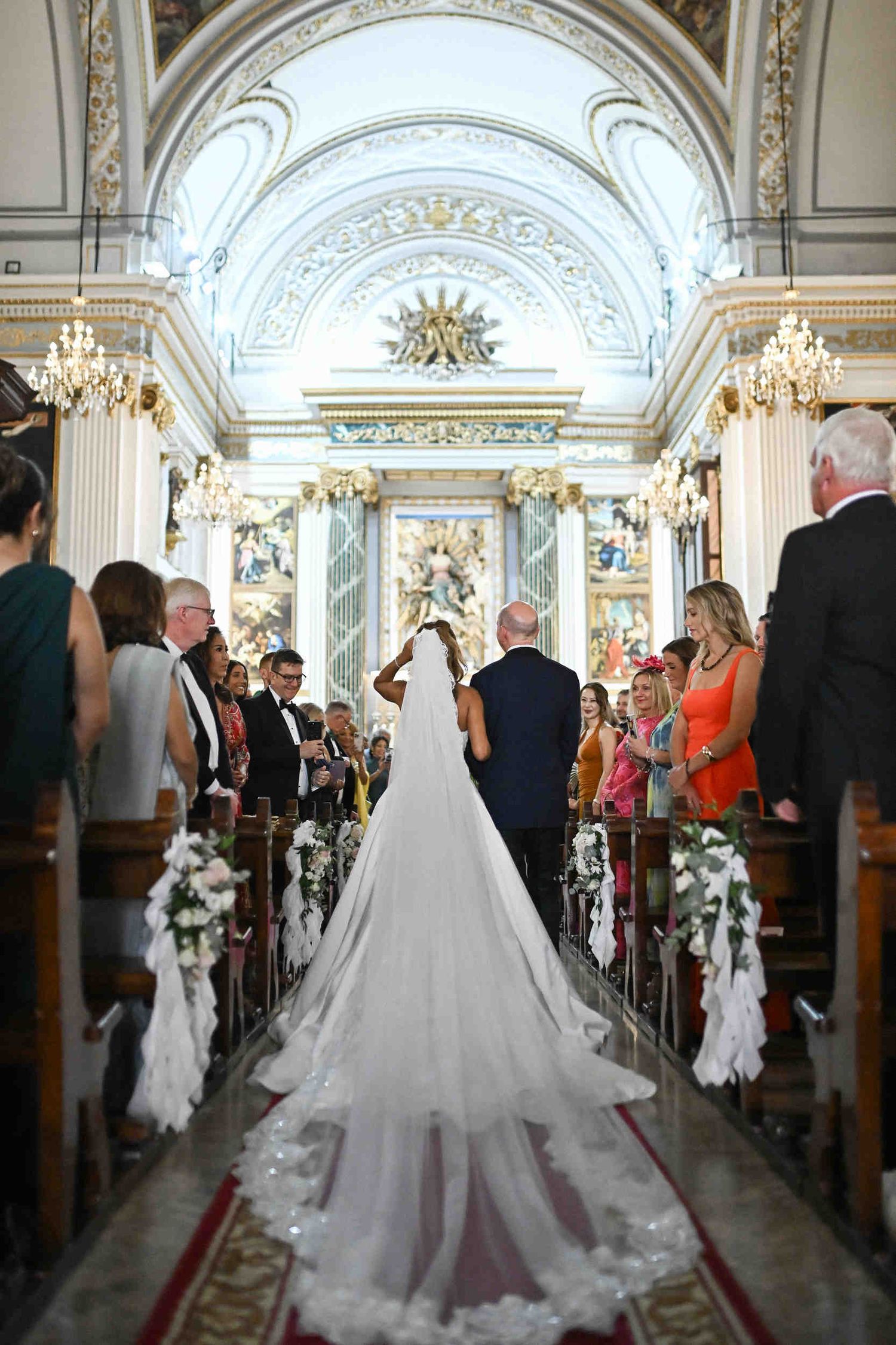 Bride walking down the aisle in a church in Valencia showing her long veil trailing along the red carpet.