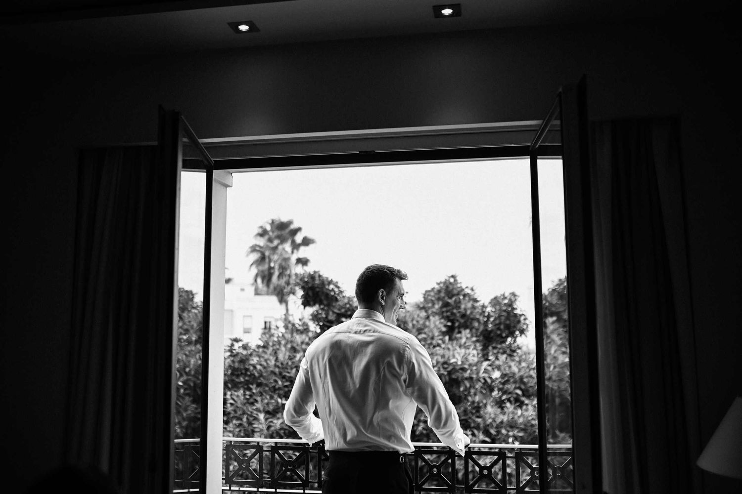 Black and white photo of a groom looking out at palm trees from a hotel room balcony at Las Arenas Resort