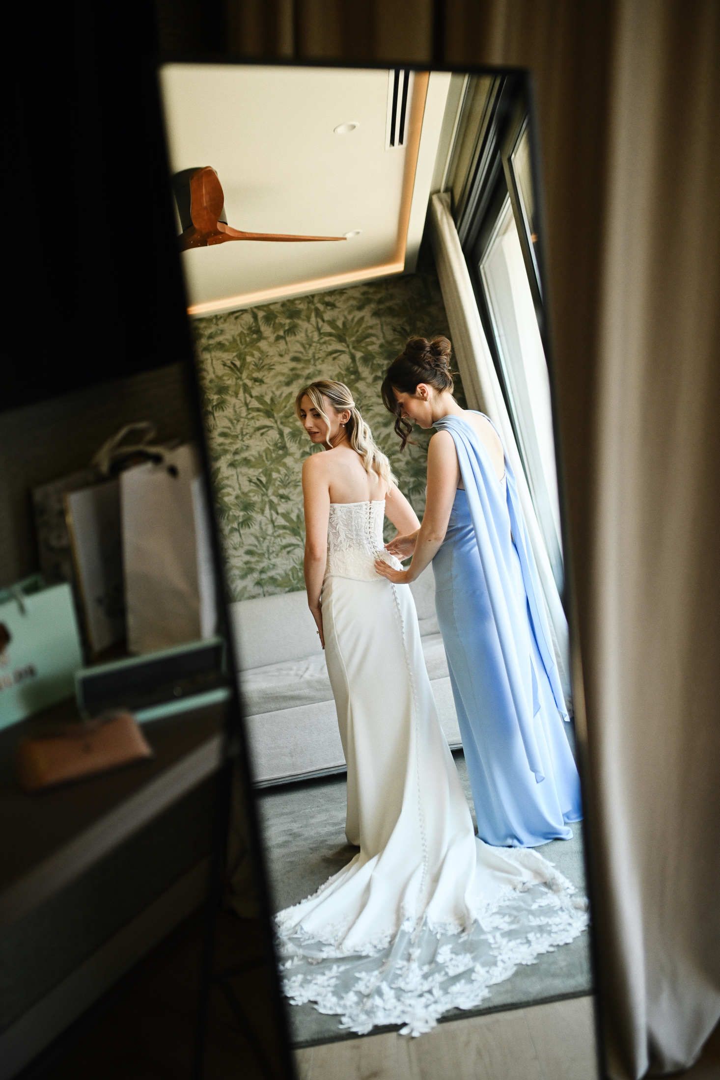 Bridesmaid helping the bride to button up her elegant wedding dress in a hotel room apartamentos Core Suite before the ceremony in Valencia.