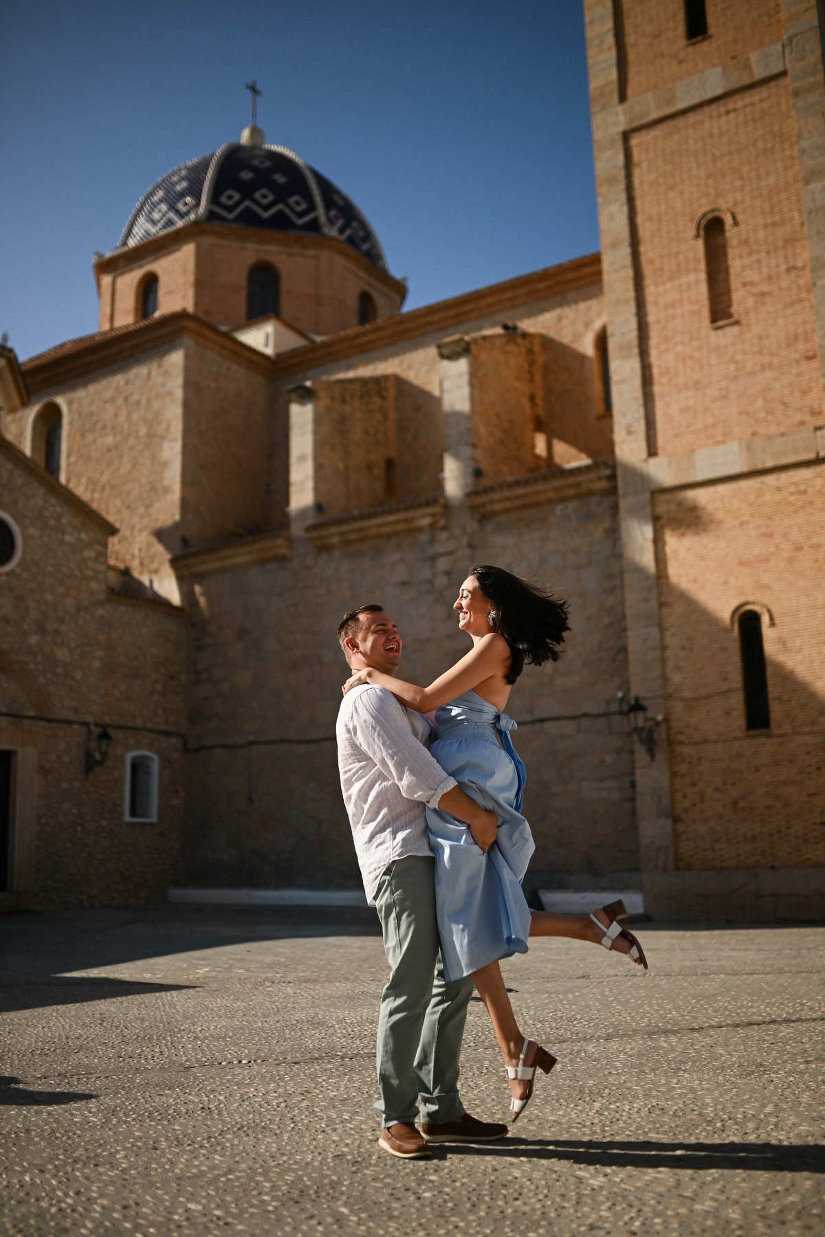 A man lifts his partner in celebration on a cobblestone square with the iconic blue domed church of Altea behind them.
