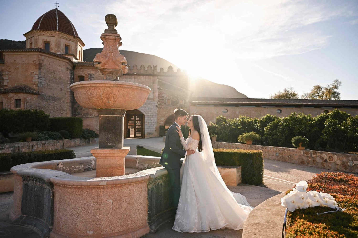 Bride and groom by the fountain in Monasterio de Santa María de la Valldigna at sunset with golden sun flare, Valencia.
