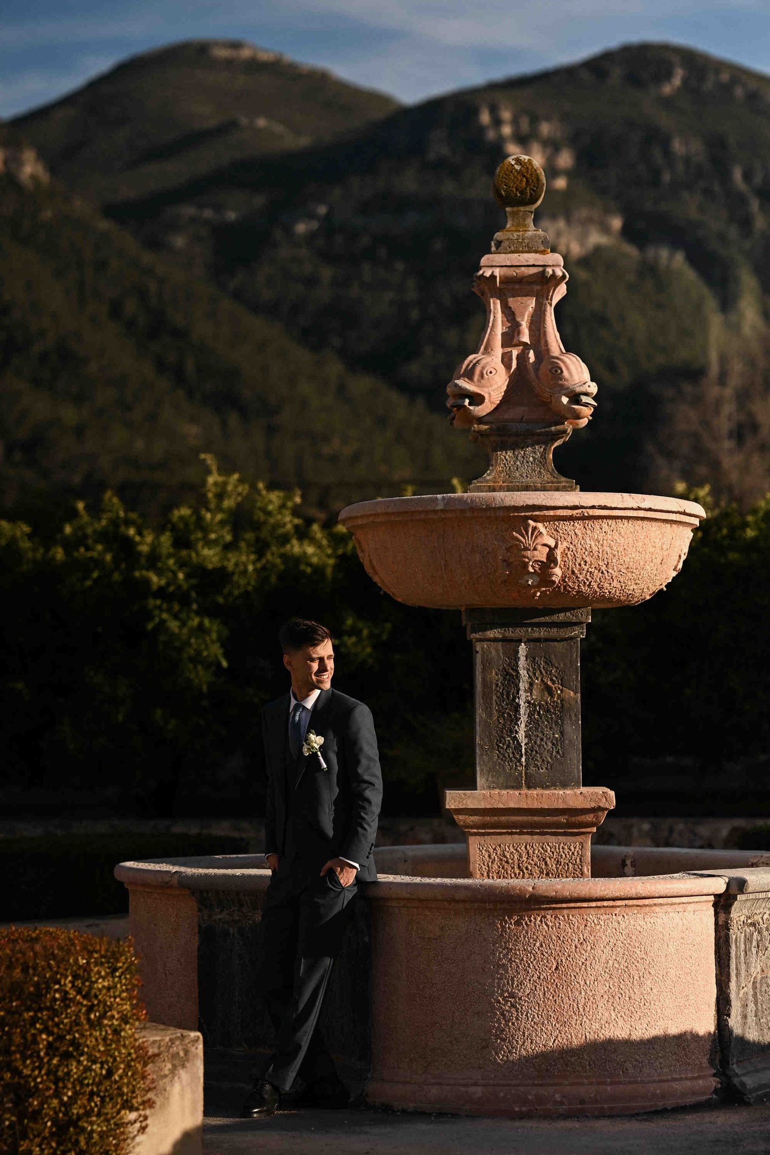 Groom standing by a fountain at sunset with Spanish mountains in the background at Monasterio de Santa María de la Valldigna.