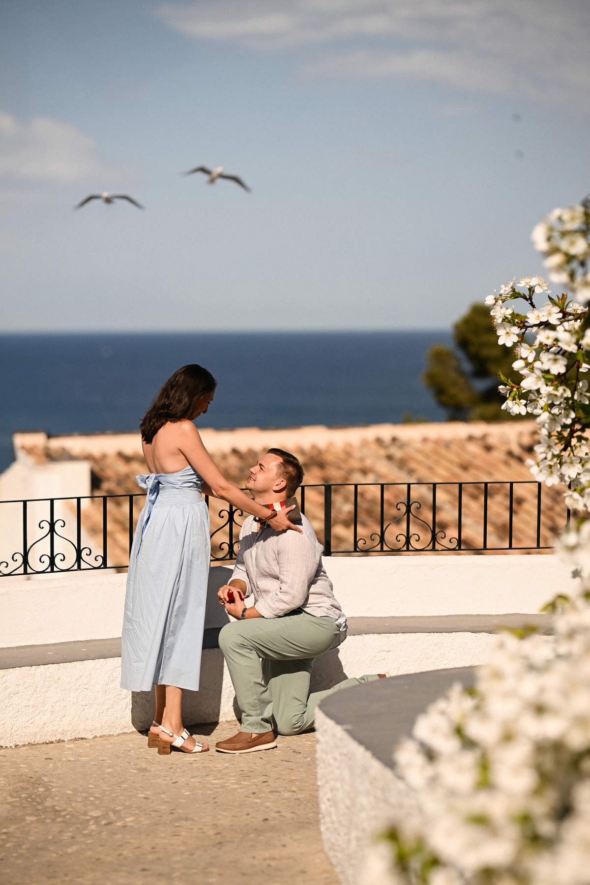 A proposal photographer in Altea captures a man kneeling with a red ring box on a sunny terrace overlooking the sea.