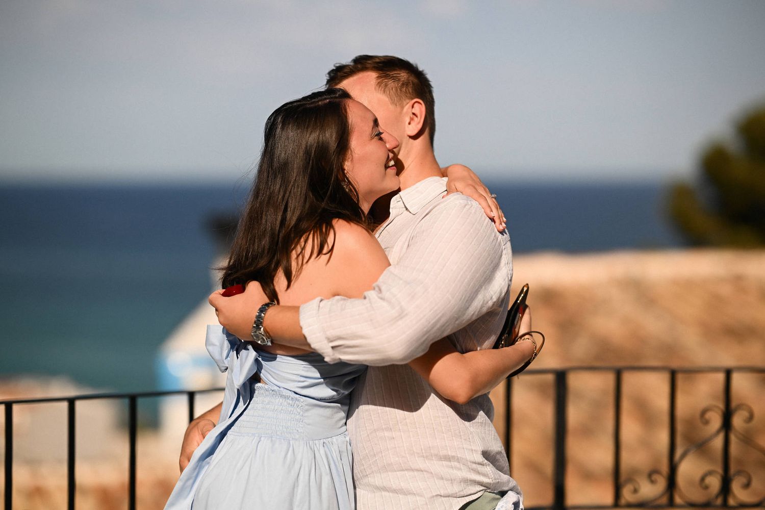 A newly engaged couple hugs tightly in the sunlight on a white balcony with the Mediterranean sea behind them in Altea.