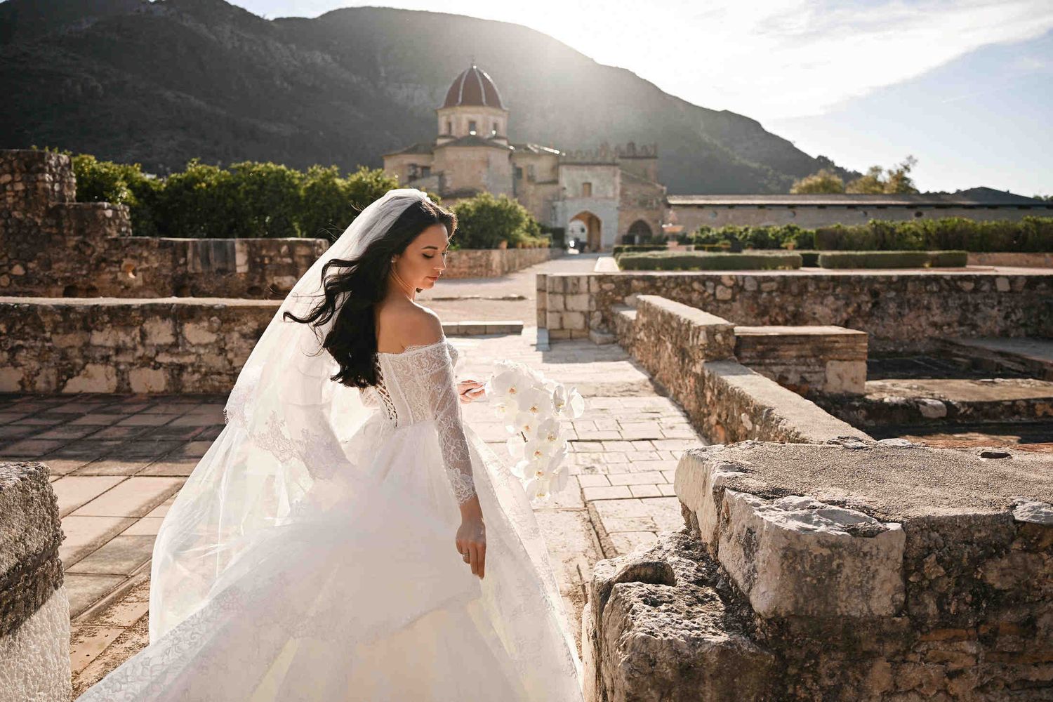 Close-up of a bride with an orchid bouquet at sunset in Monasterio de Santa María de la Valldigna, hair and veil flowing in the breeze.