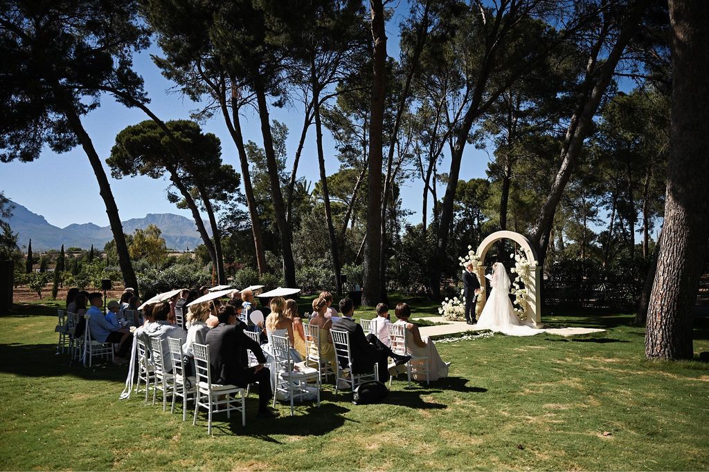 Beautiful outdoor wedding ceremony setup on a sunny day with guests seated on the grass and mountains in the background.