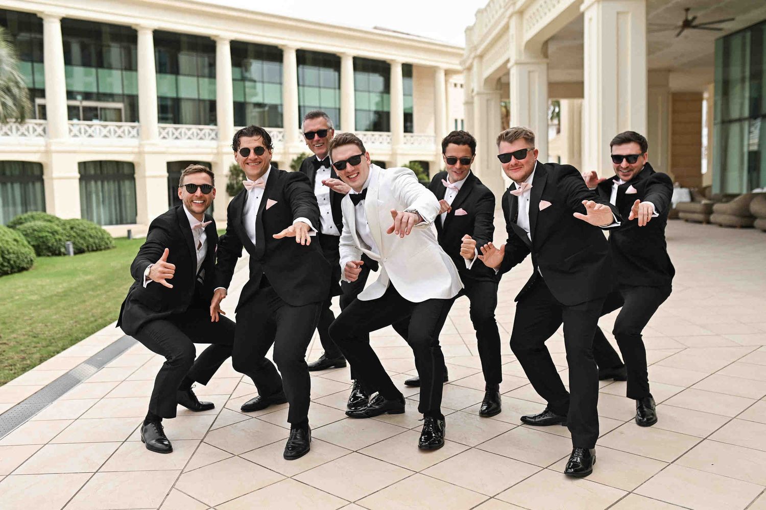 Groom and groomsmen wearing black tuxedos and sunglasses posing playfully near Hotel Las Arenas colonnade