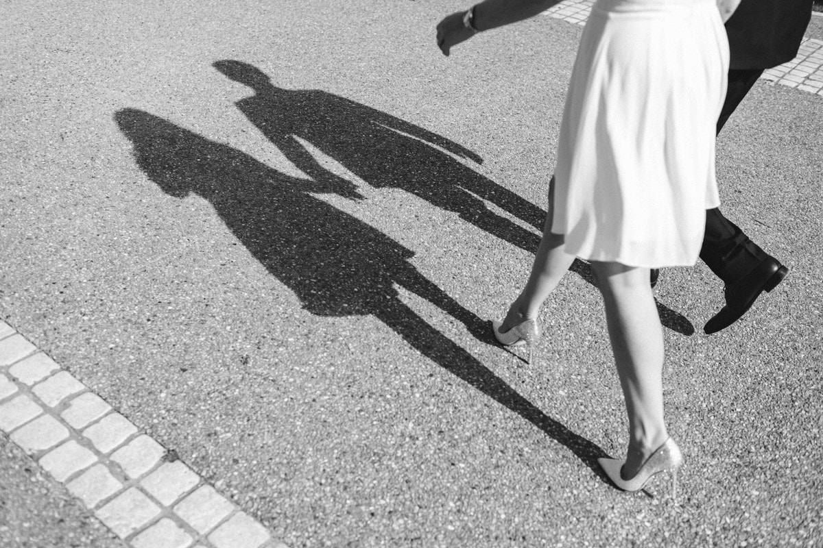 Black and white view focusing on the long shadows of a couple holding hands and walking on a paved Paris street