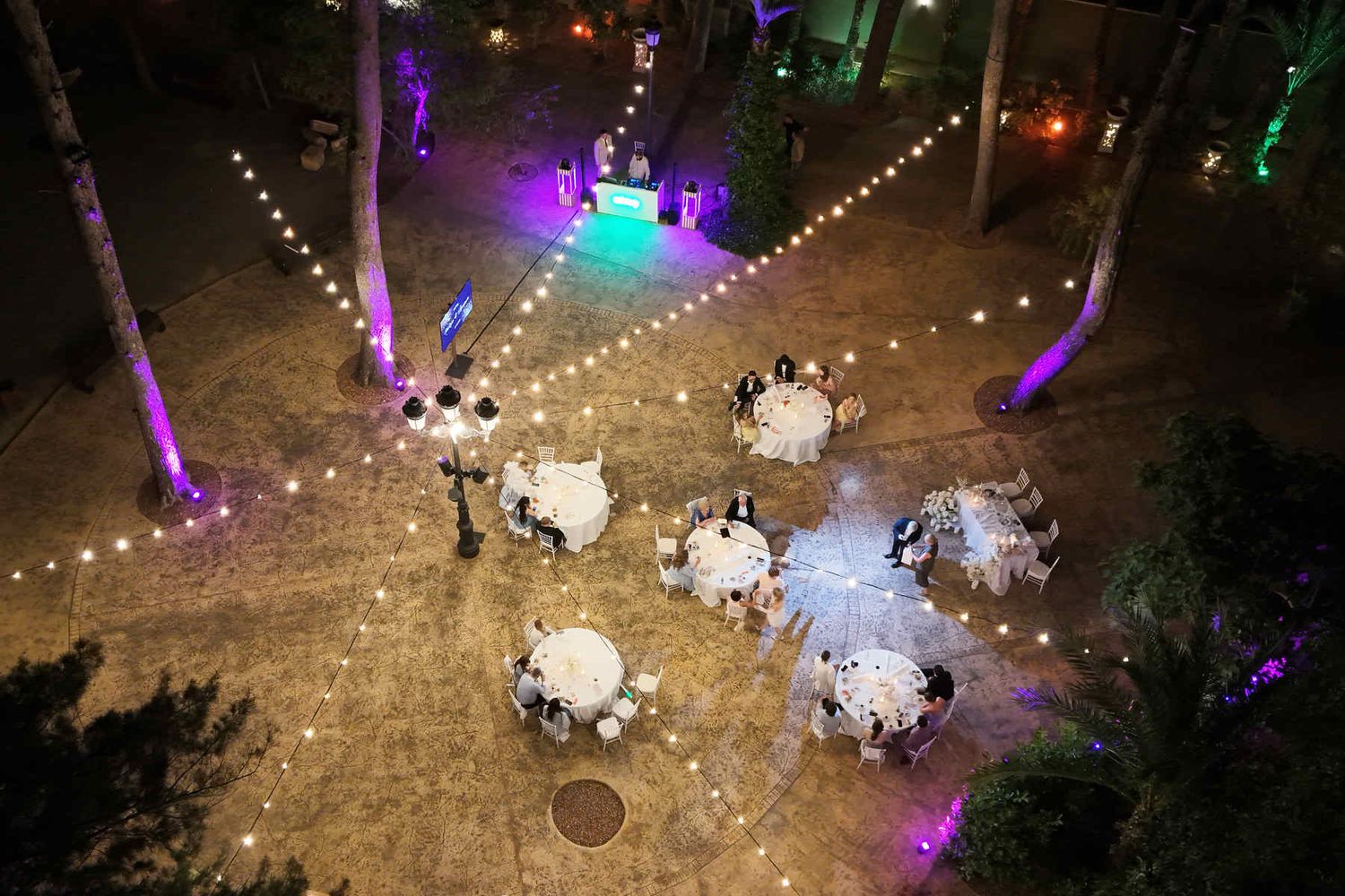 High-angle drone shot of an outdoor wedding reception party at night featuring round tables, warm string lights, and a DJ dance floor.