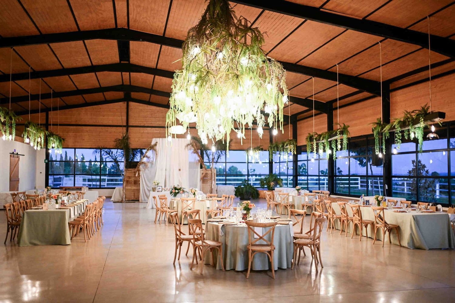 Large indoor wedding reception hall featuring a massive hanging greenery chandelier with warm light bulbs.