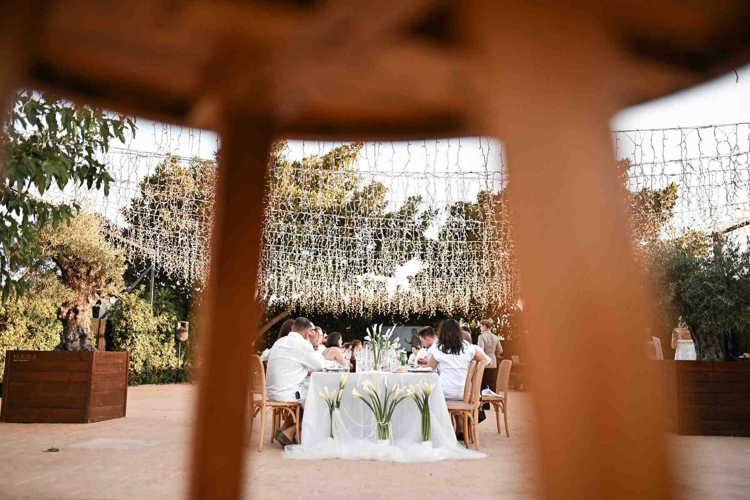 Creative blurred foreground framing of an outdoor wedding banquet dinner with guests in white attire.