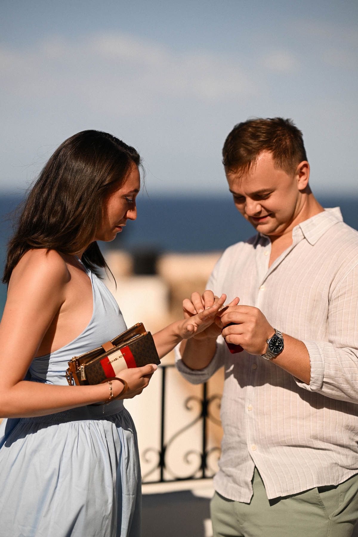 A man slides an engagement ring onto a woman's finger on a white terrace overlooking the ocean after proposing in Altea.