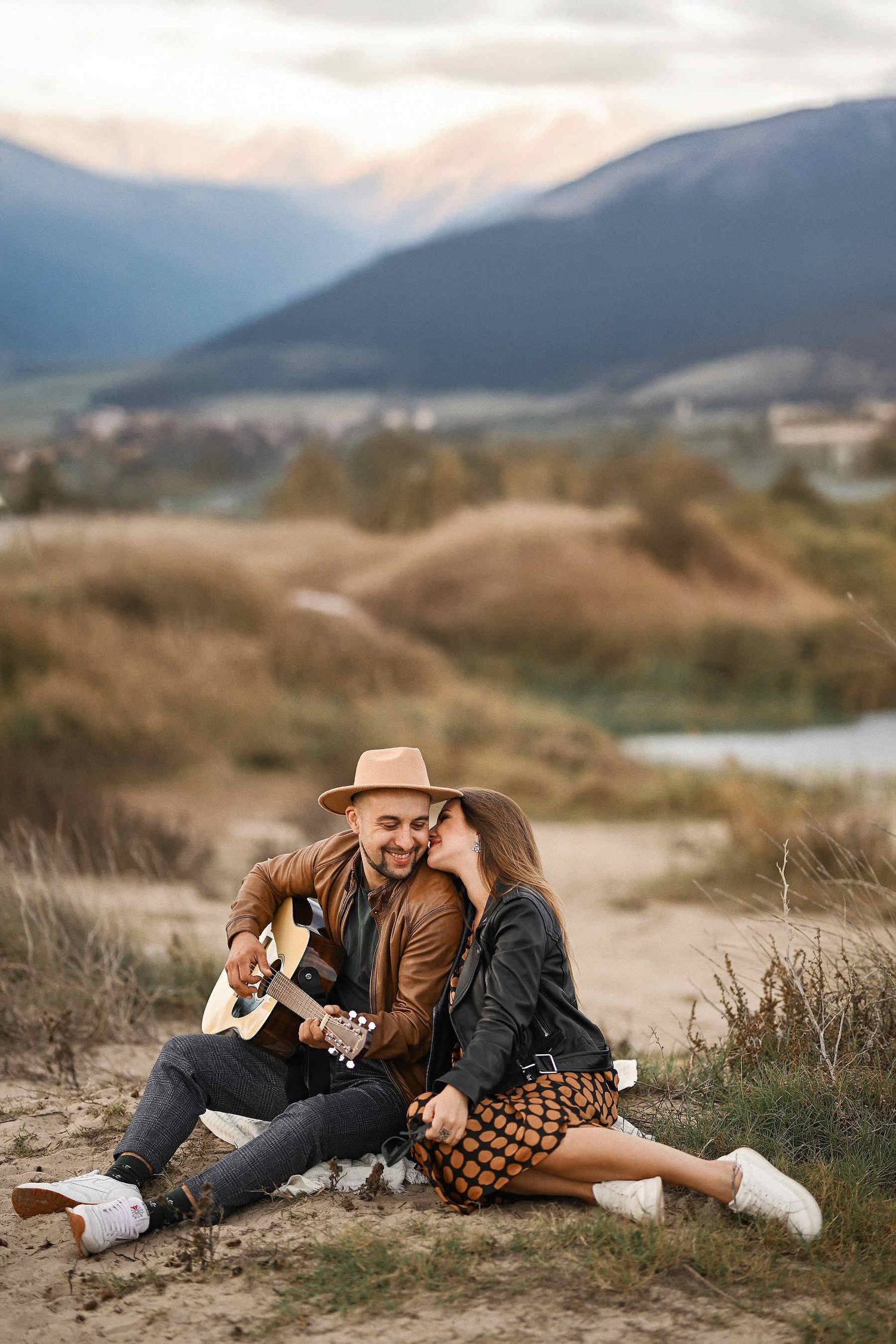 Coastal landscape of beach grass and mountains while the groom plays guitar during an engagement photoshoot in Spain