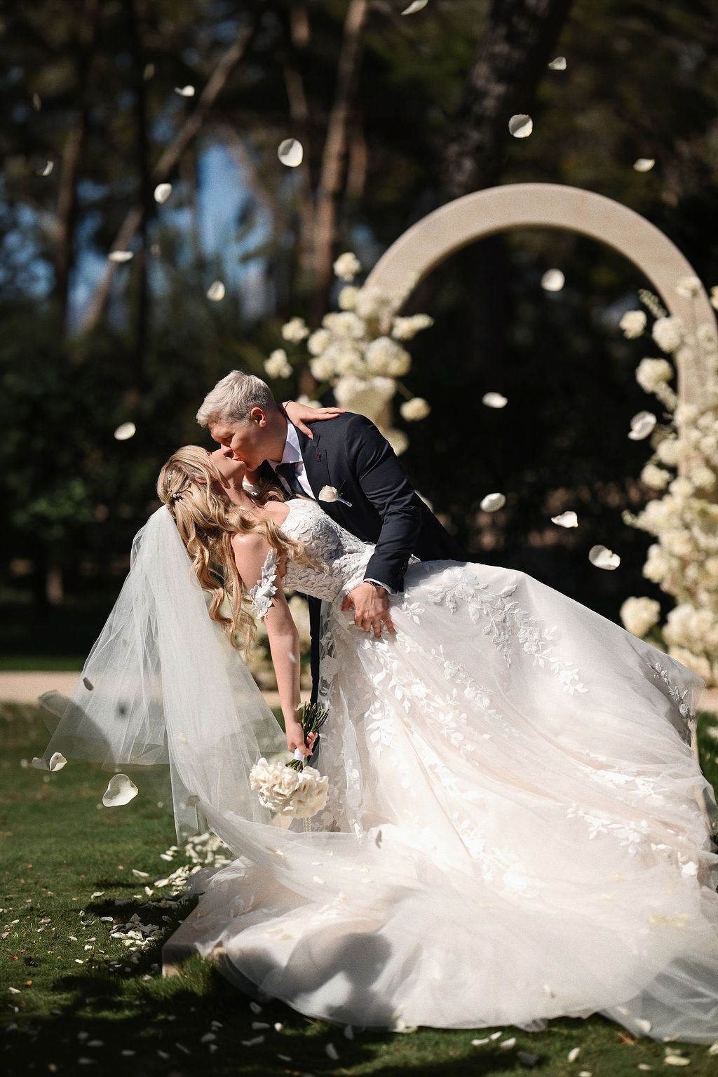 Groom dips and kisses the bride under the historic stone arch at El Bosque ceremony site, during a rose petal toss in Benidorm, Spain.