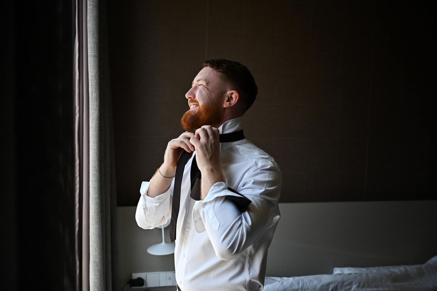 Smiling groom fixing his tie while getting ready for the wedding ceremony in a hotel room during morning prep.