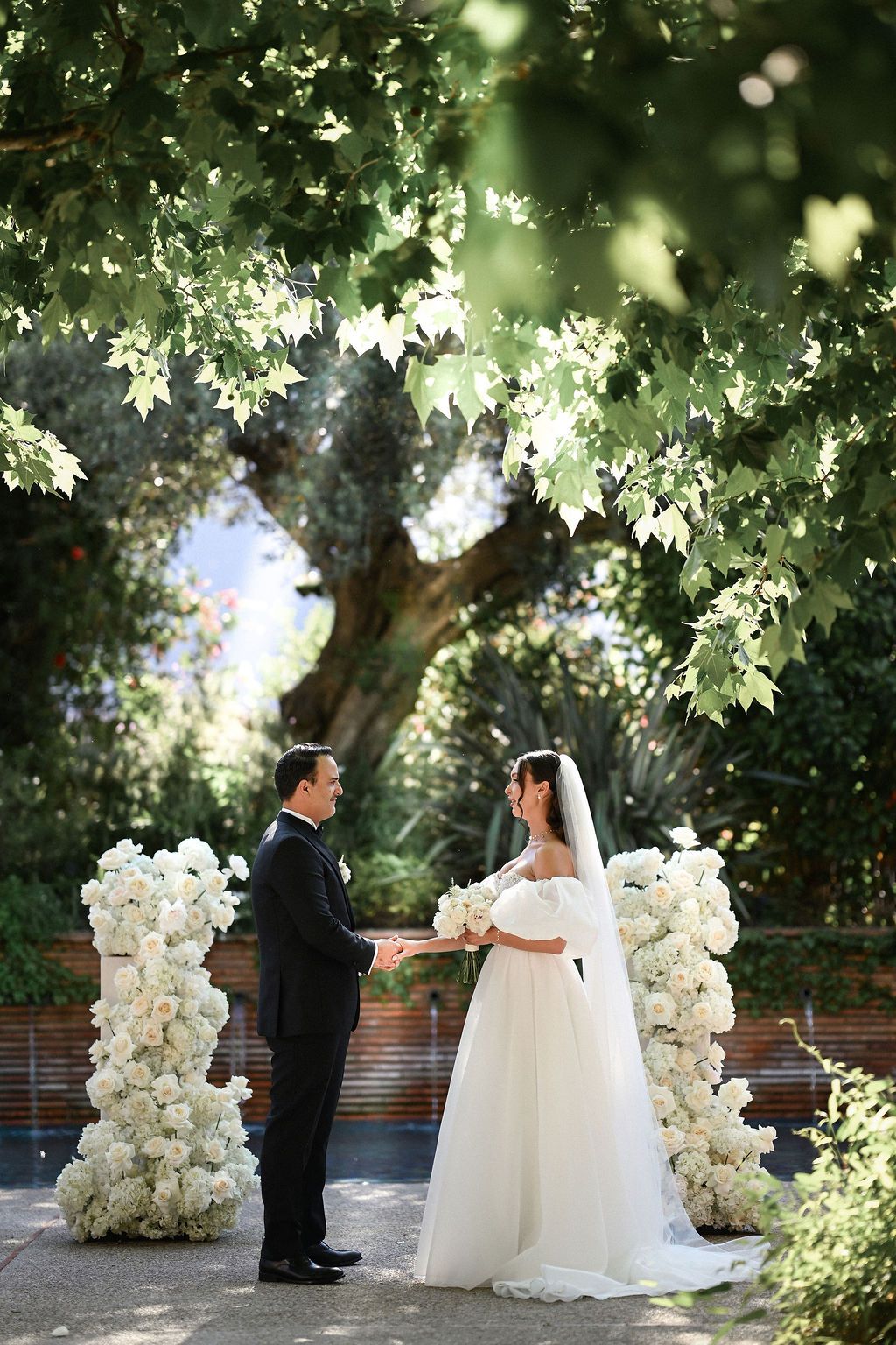 The newlyweds holding hands by the floral arch in the warm golden hour sunlight. Touching wedding ceremony photography by Darista.