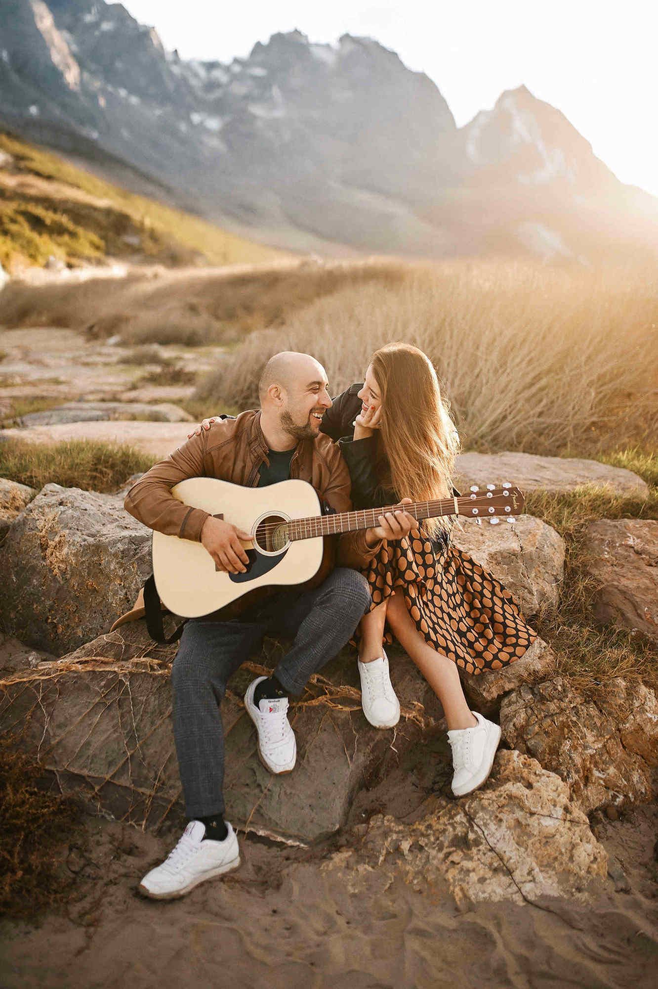 Engagement photographer Spain. Couple sitting on seaside rocks with a guitar against a backdrop of Spanish mountains and blue water.