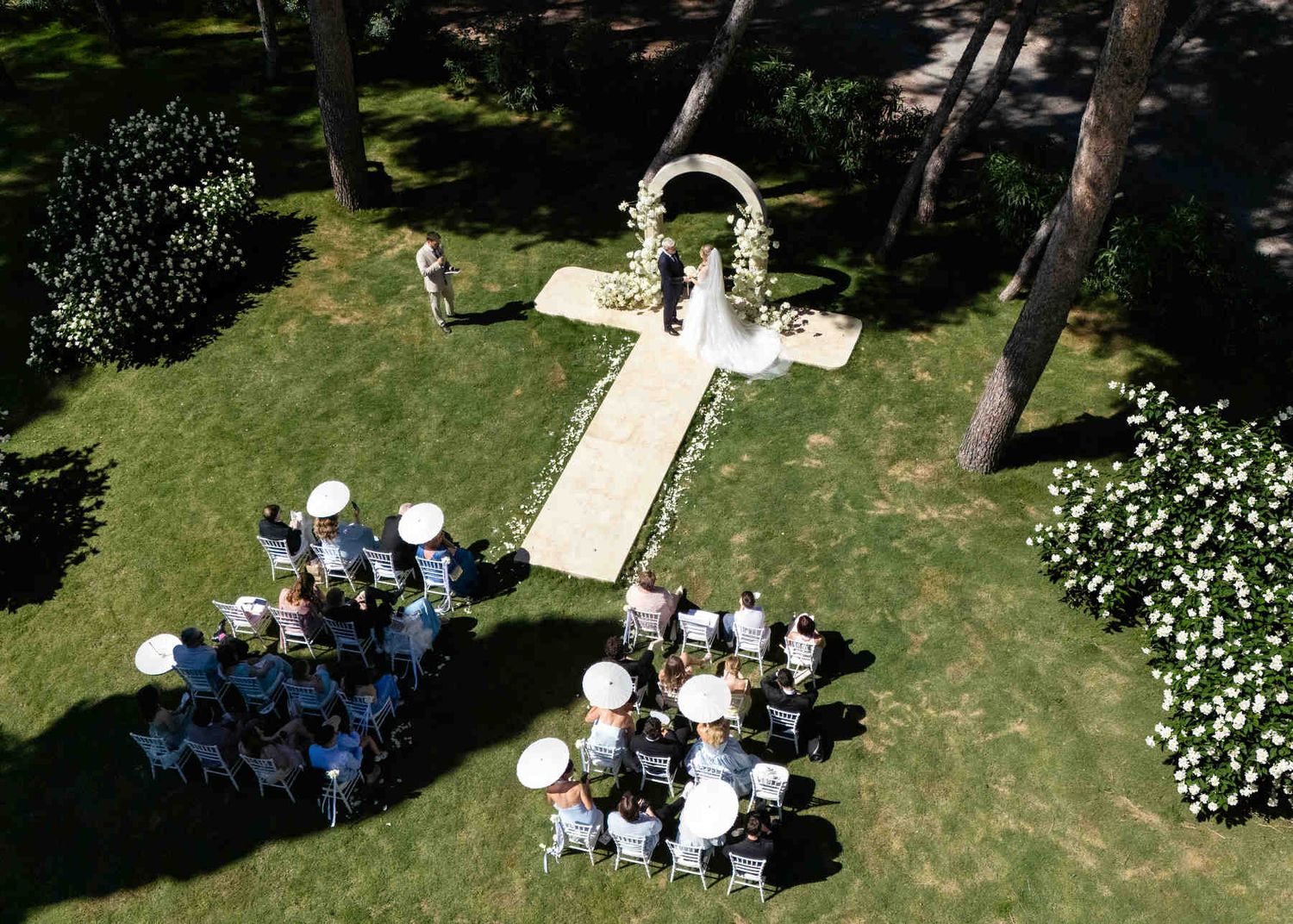 Aerial drone photograph of an outdoor wedding ceremony in Spain with a white floral arch and guests holding parasols on a green lawn.