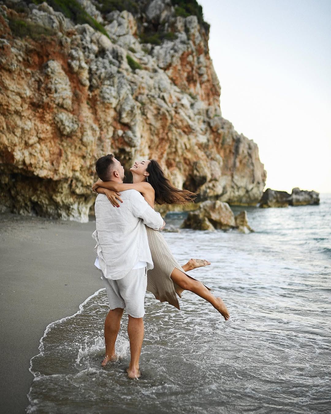 Man lifting his partner by the water at a hidden rocky beach south of Valencia during a wild and romantic engagement photoshoot.