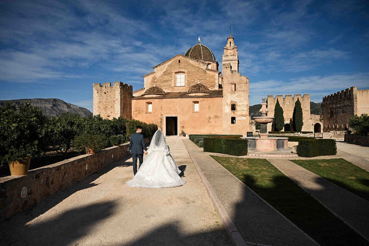 Bride and groom walking in the historic courtyard of Monasterio de Santa María de la Valldigna. Wide shot of Valencian heritage architecture