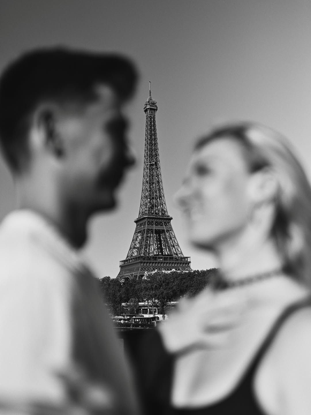 Black and white engagement portrait framing the Eiffel Tower in sharp focus between the blurred silhouettes of the couple.