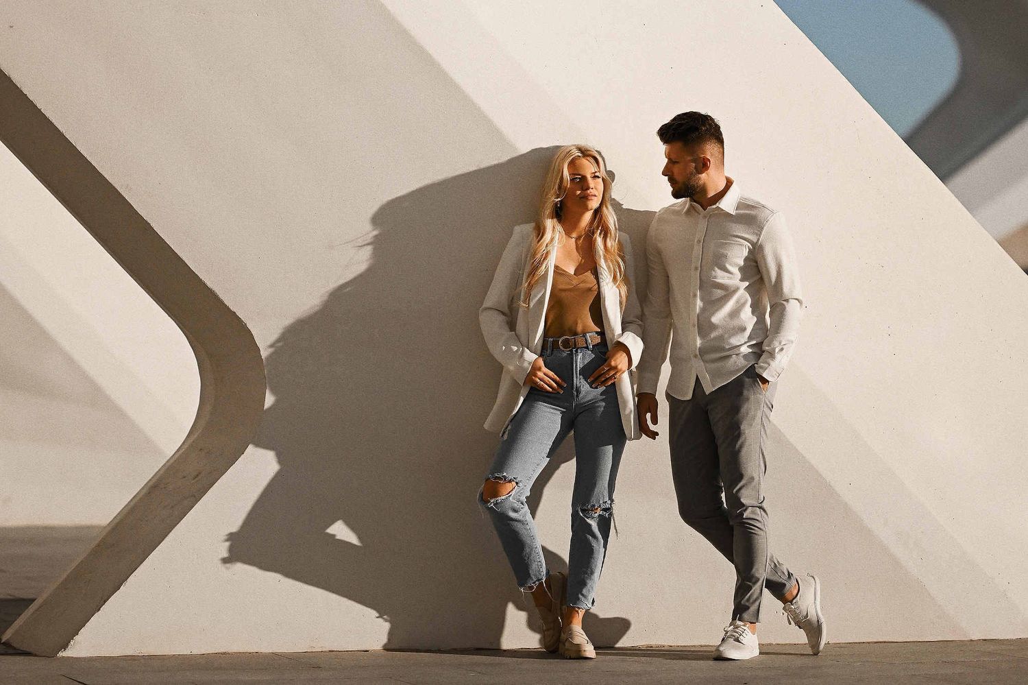Pre-wedding photo session Spain. Couple in chic white blazers and jeans posing against minimalist white architecture in a Spanish town.