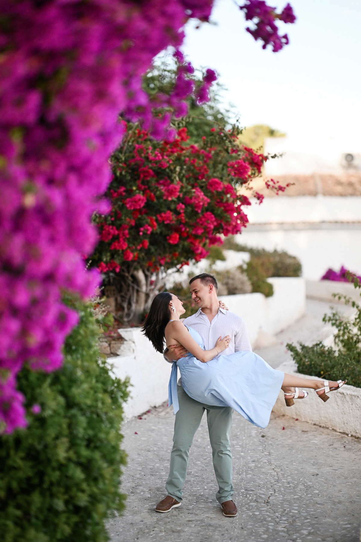 A man happily carries his partner in his arms past vibrant pink bougainvillea bushes during an Altea proposal session.