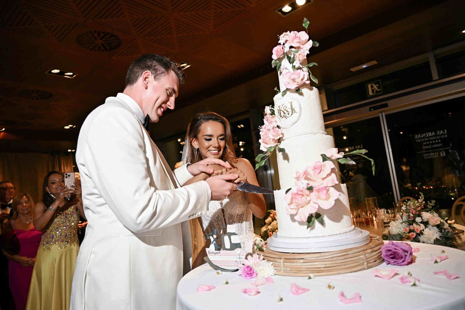 Bride and groom cutting a four-tier white wedding cake decorated with fresh pink roses during reception.