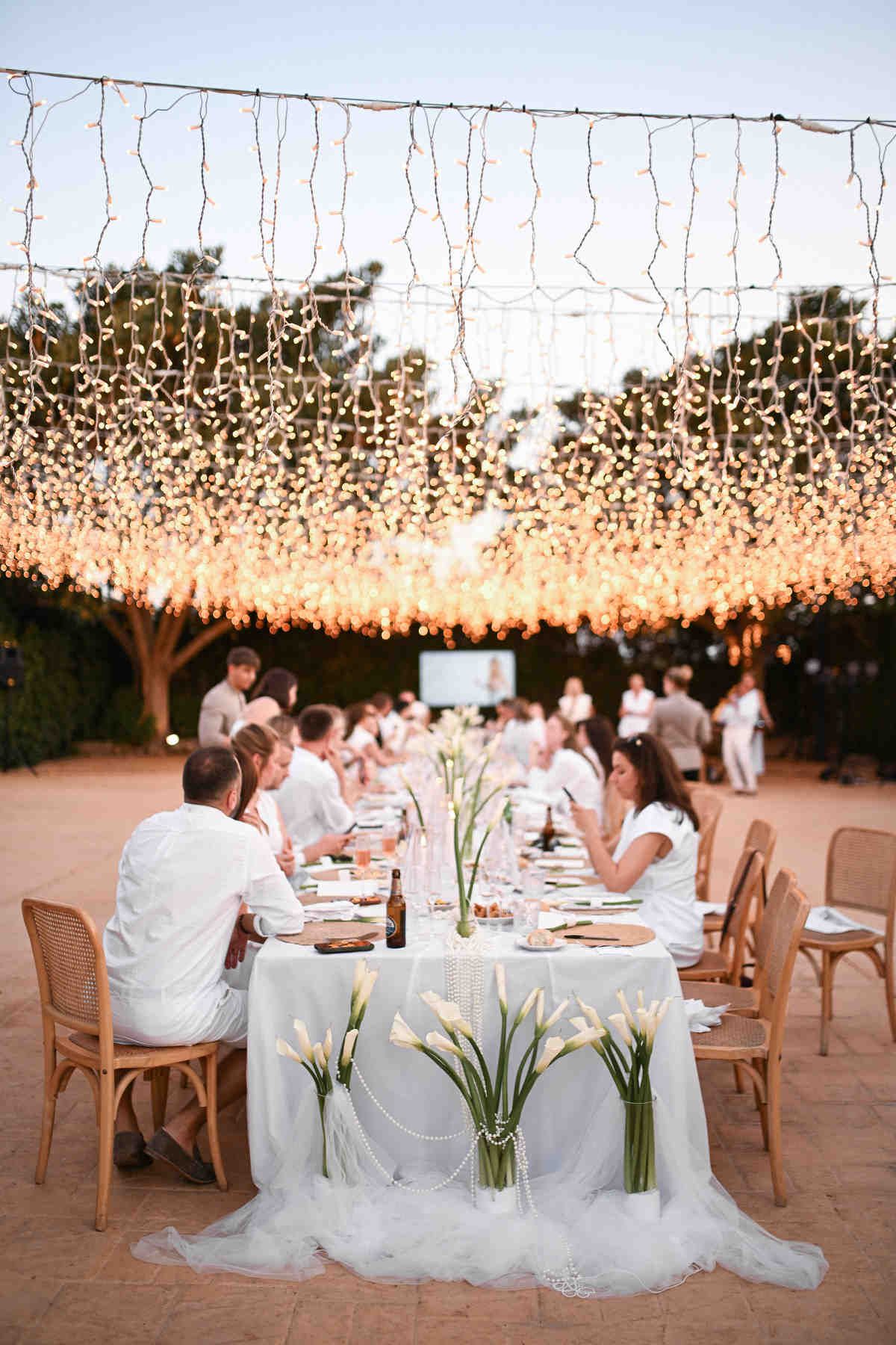 Outdoor wedding dinner with guests seated at a long table decorated with calla lilies under warm string lights.
