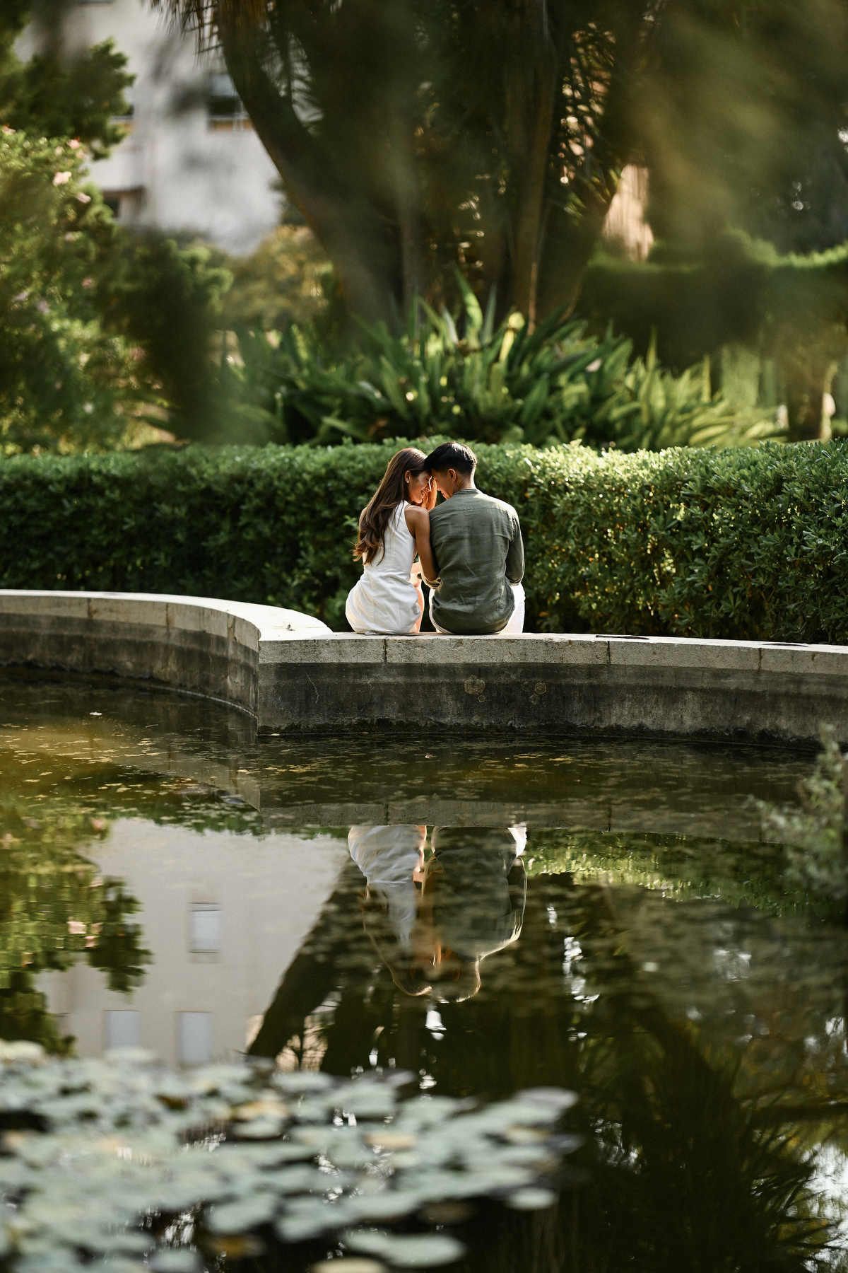 A couple sits on a stone wall by a pond, touching foreheads, with their clear reflection visible in the water below.
