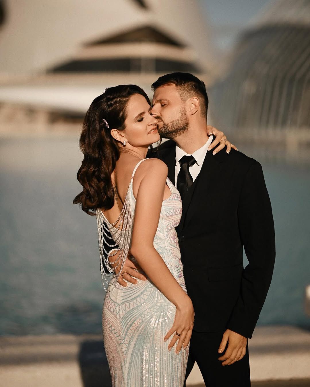 A groom kisses his bride's cheek beside a blue pool, framed by the white curves of Valencia's City of Arts and Sciences.
