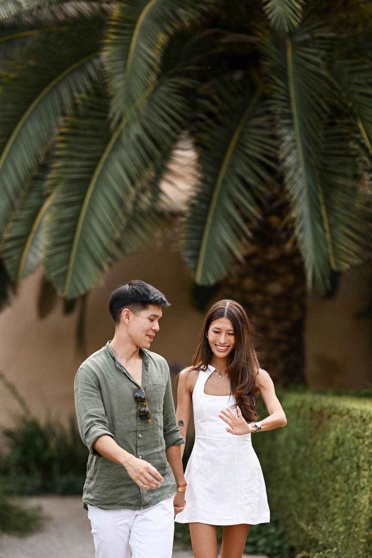 A smiling couple walking hand in hand under large palm tree leaves after a surprise marriage in a neoclassical park.