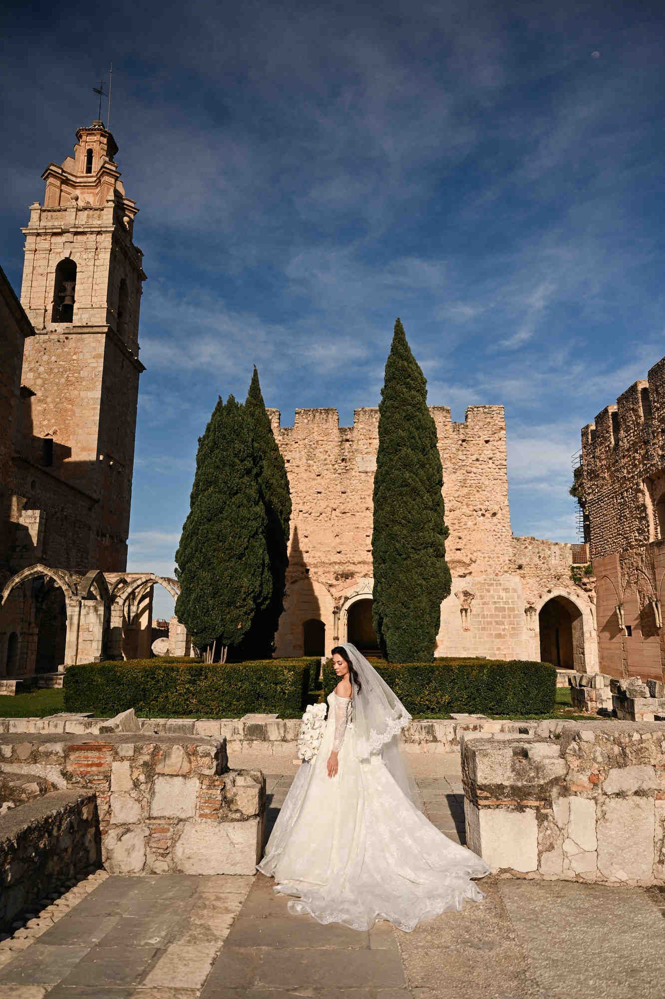 Full-length shot of a bride in a wedding dress against cypresses and historic tile roofs of Monasterio de Santa María de la Valldigna.