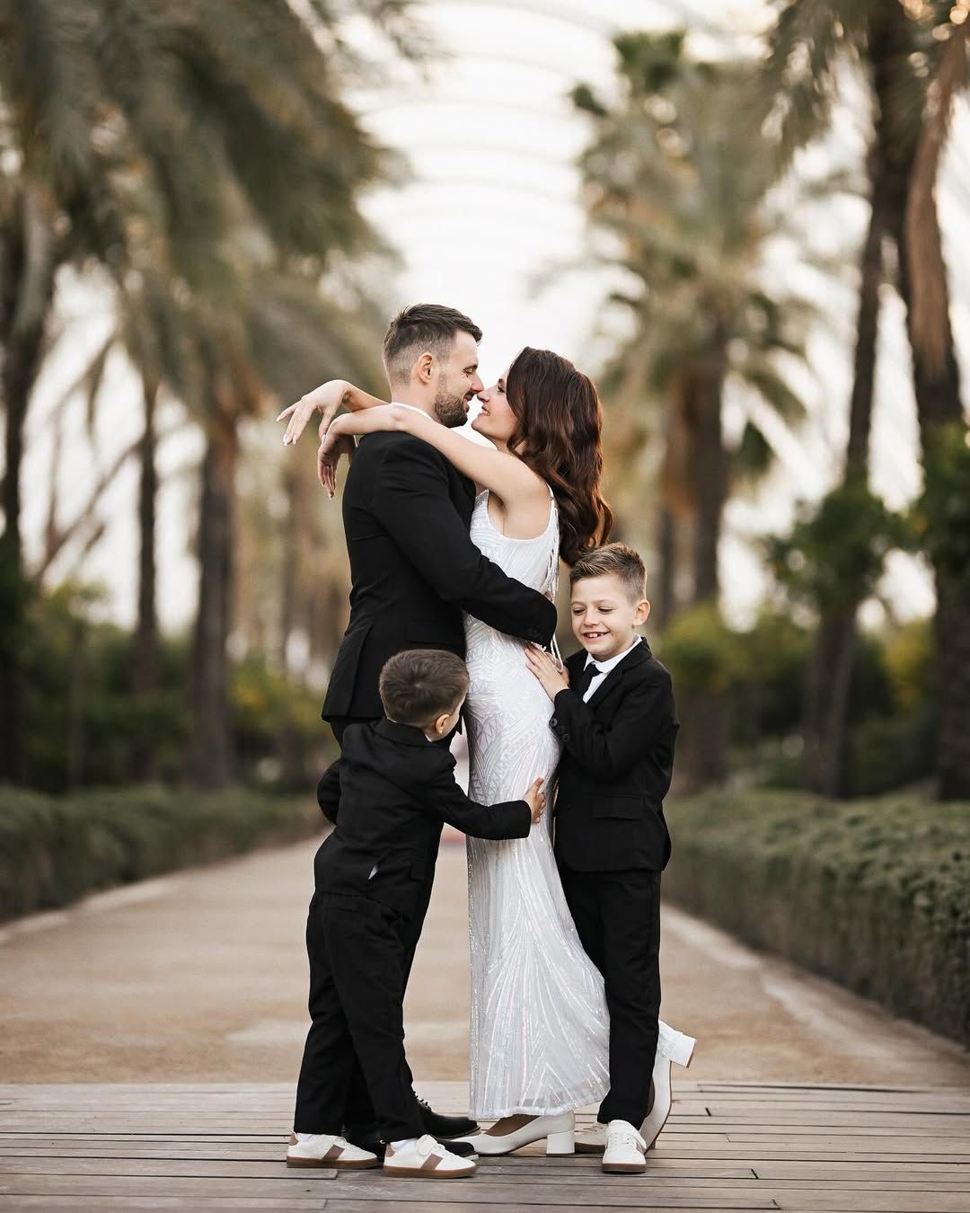 A bride and groom embrace in Valencia while their two young sons in black suits playfully hug them on a palm-lined path