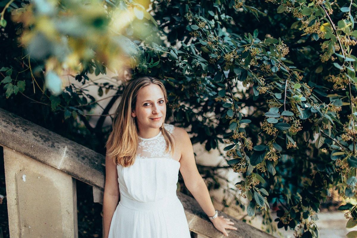 A bride-to-be in a white dress leans against a stone balustrade surrounded by lush green foliage near the Seine river.
