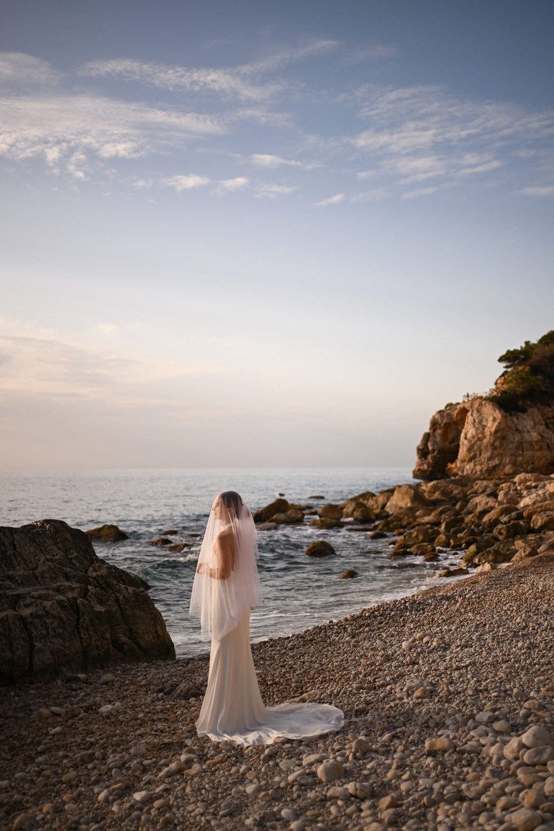 Bride in a white gown and veil covering her face standing on a pebble shore near cliffs at dusk.