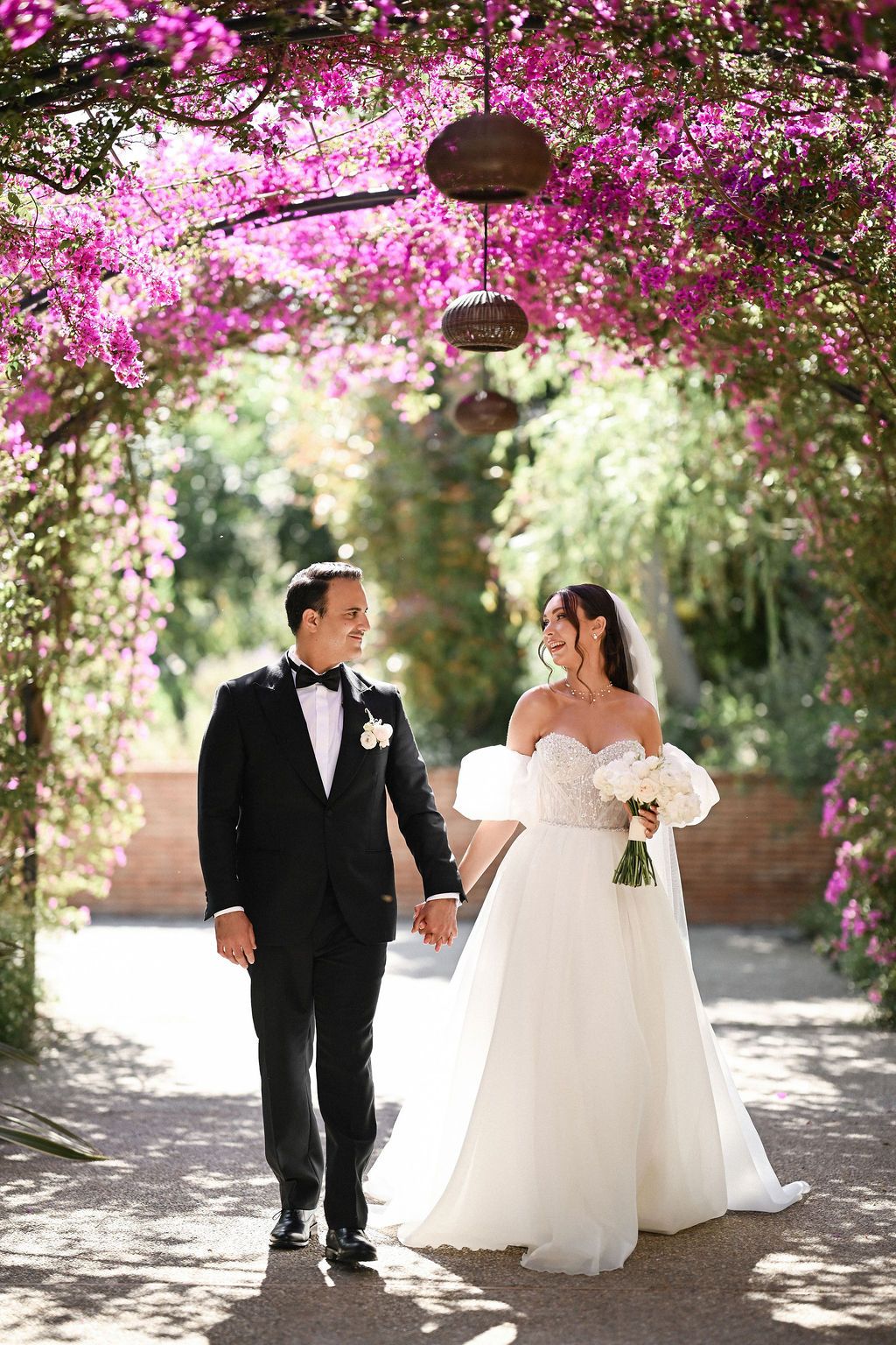 The newlyweds walking along a path of vibrant blooming pink flowers in the lush Mediterranean gardens of Finca del Canónigo villa.