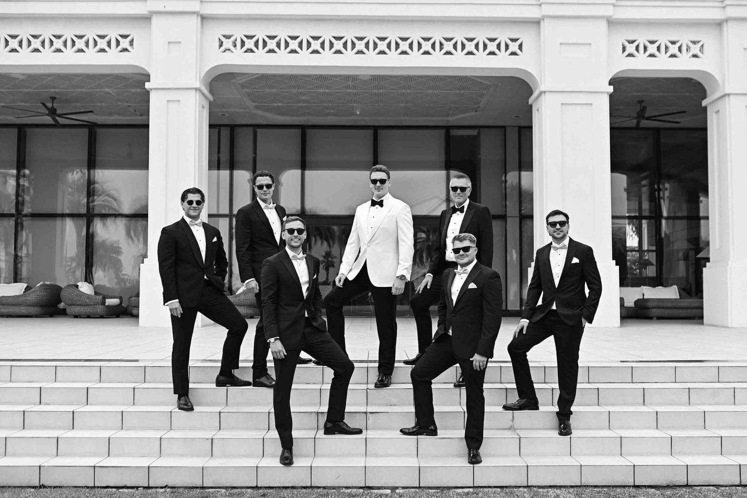 Black and white group photo of groom and groomsmen in black tuxedos posing on hotel entrance stairs.