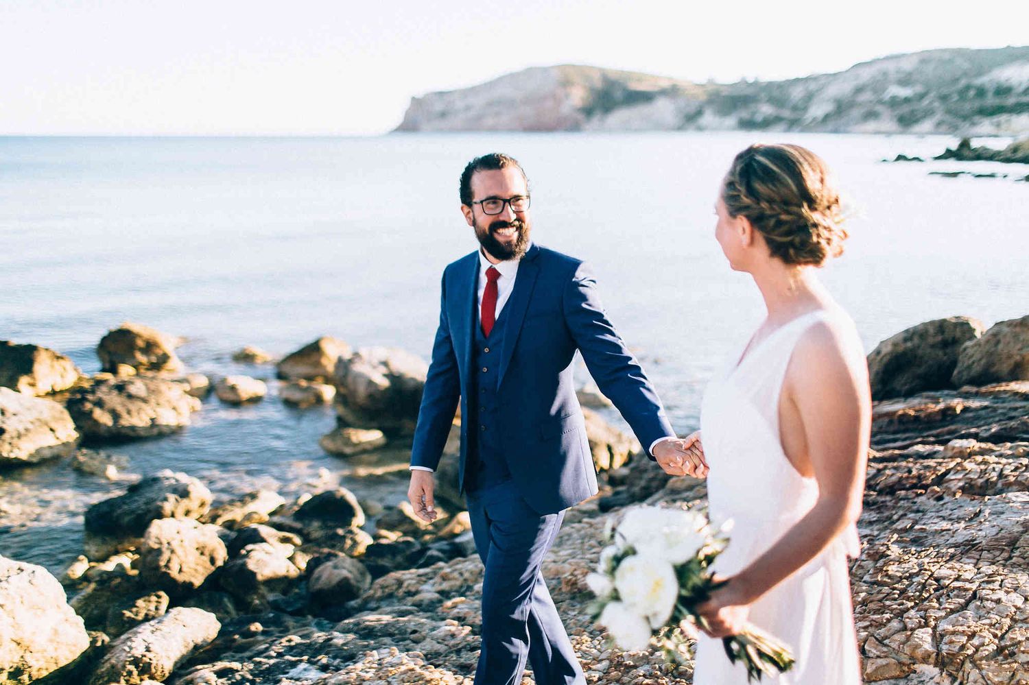 Happy wedding couple strolling along the scenic European coast in Greece, smiling and looking at each other amidst breathtaking sea views.