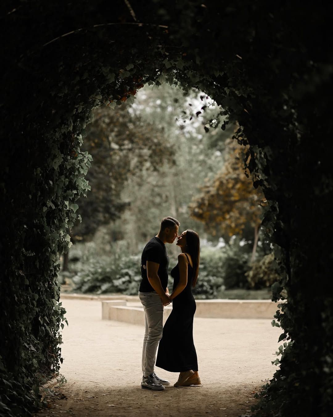 A couple holds hands and leans in for a kiss under a dark green ivy archway during a love story session in Valencia.