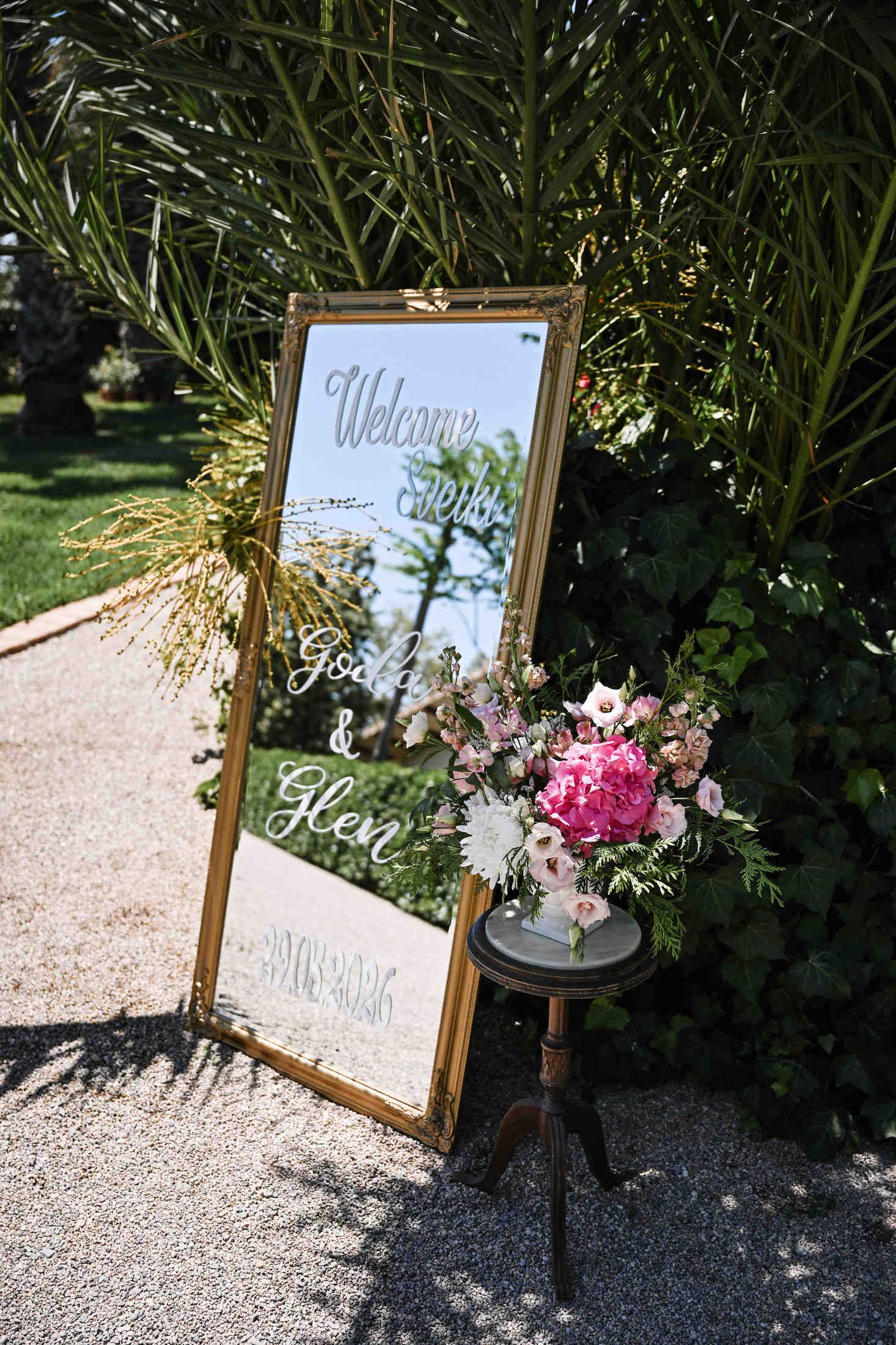 Elegant mirror wedding welcome sign decorated with fresh flowers at the entrance of Villa Dela venue in Valencia.