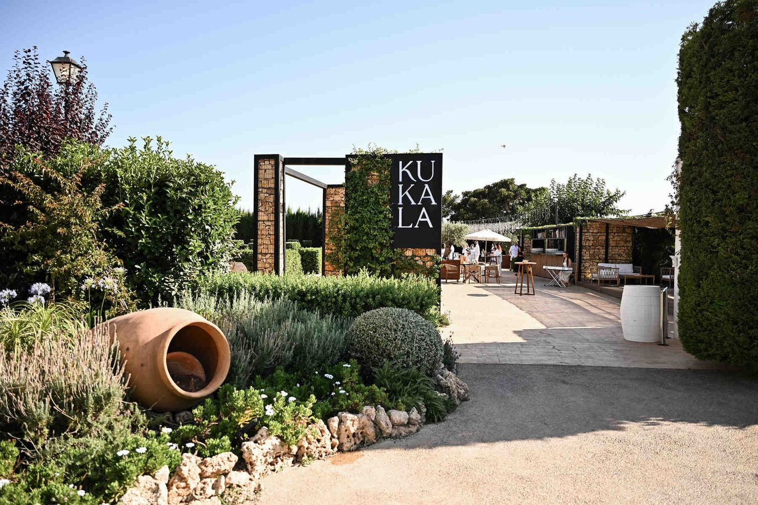 Main entrance of Alquería Kukala wedding venue in Valencia featuring a black sign, large terracotta pot, and lush green landscaping.